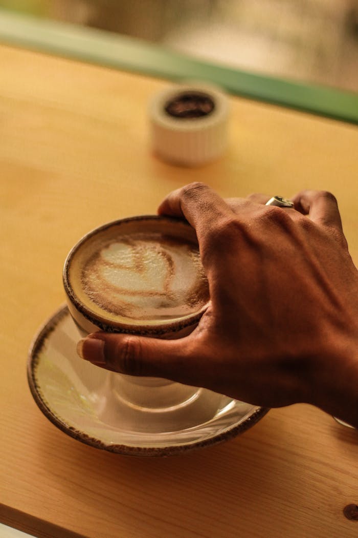 Close-up of a hand holding a latte with foam art on a saucer at a cozy cafe table.